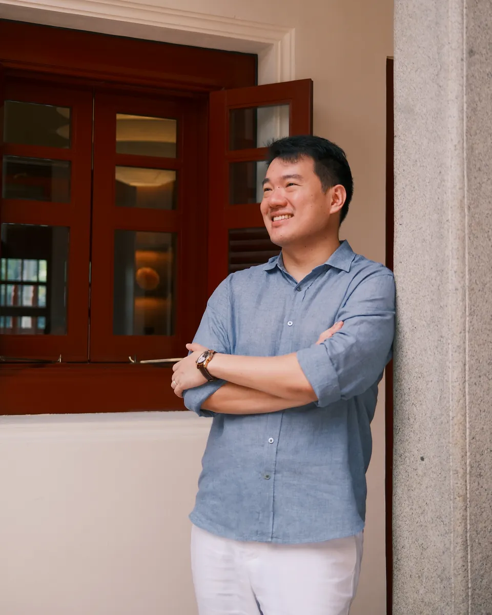 Sean Chan, arms crossed and smiling, leaning against a stone pillar beside a shophouse's red lacquered windows